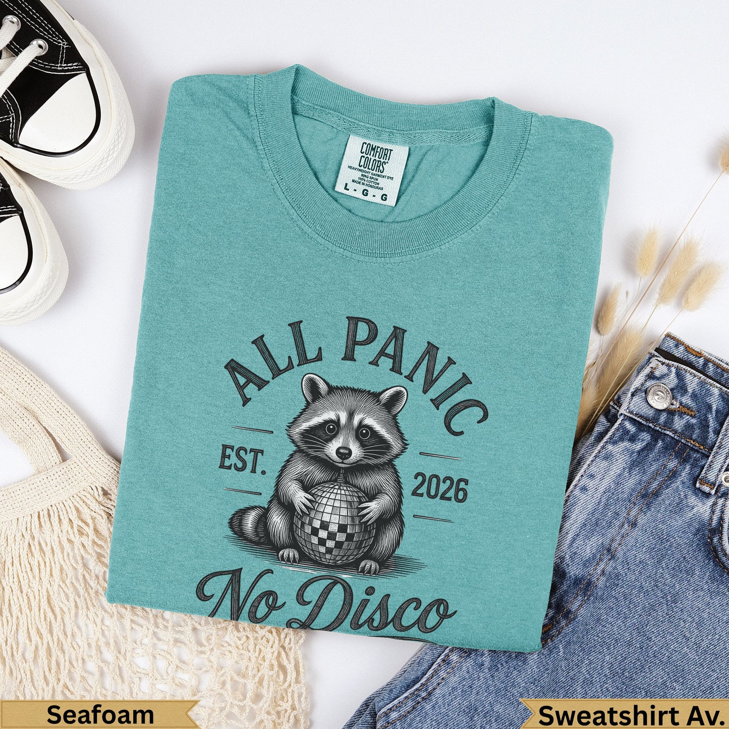 Teal t-shirt with raccoon holding disco ball, "All Panic Est. 2026 No Disco" text.