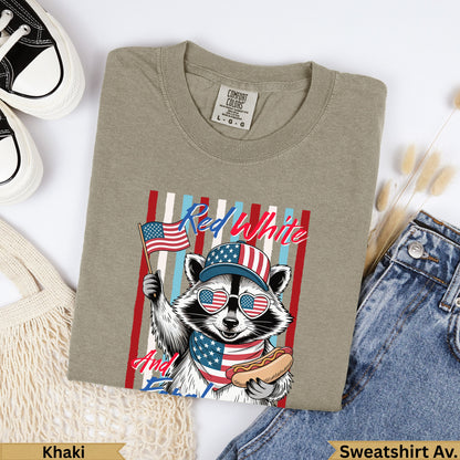 Raccoon wearing a patriotic hat and bandana holding an American flag and hot dog.