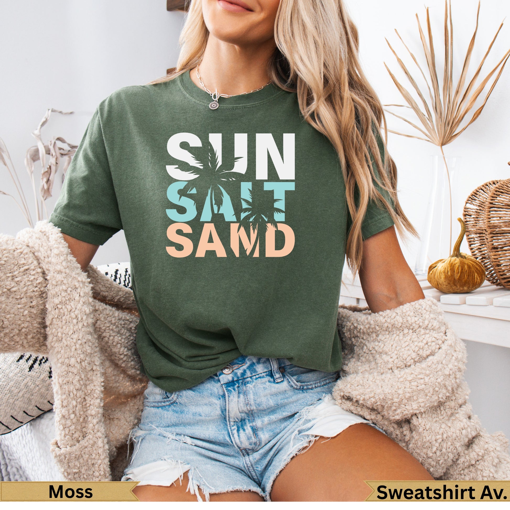 Green t-shirt with "SUN SALT SAND" text, woman wearing it with denim shorts.
