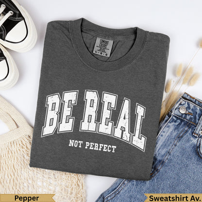 Dark gray t-shirt with "BE REAL NOT PERFECT" text, beside jeans.