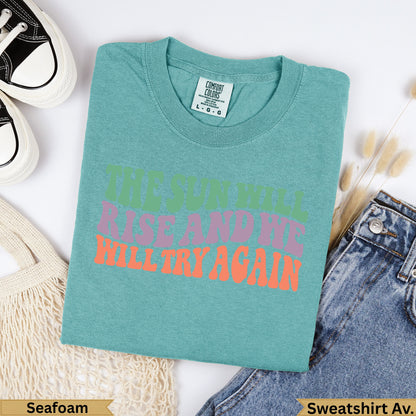 Teal t-shirt with "The sun will rise and we will try again" text.