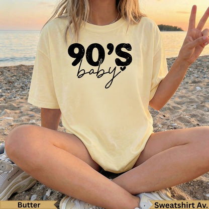 Woman in butter yellow t-shirt with "90's baby" text, sitting on beach sand.