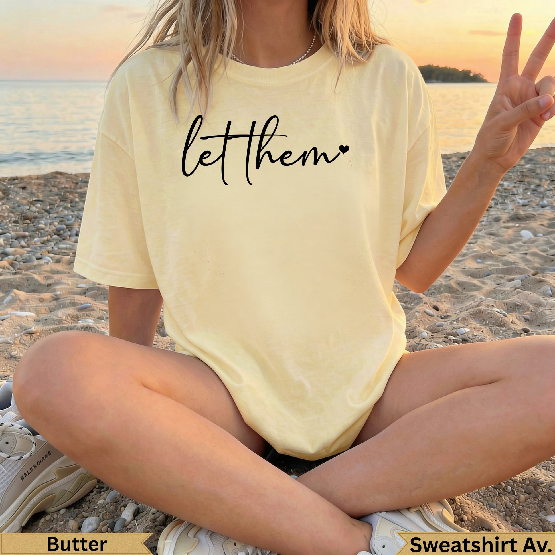 Woman in yellow sweatshirt with "let them" text, sitting on beach sand.