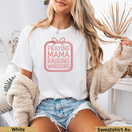 White t-shirt with "Praying Mama Raising Warriors" text design in pink, worn by a woman.