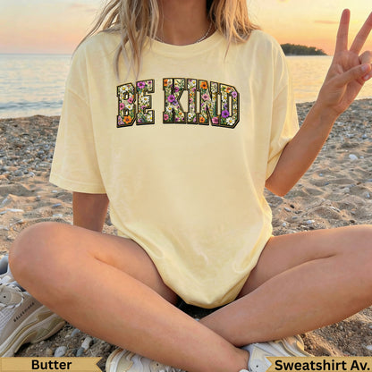 Yellow "BE KIND" graphic tee with floral letters, worn by a person on a beach.