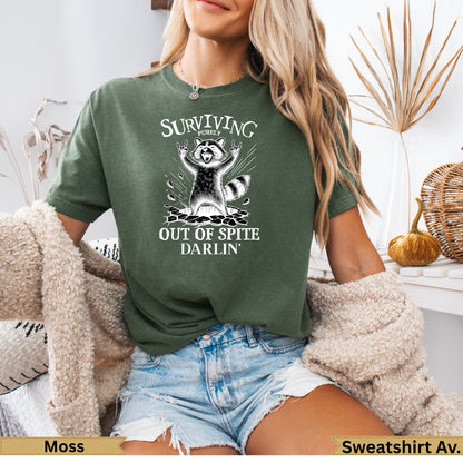 Green t-shirt with raccoon graphic and text "Surviving Purely Out of Spite Darlin'".
