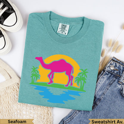 Teal t-shirt with pink camel, yellow sun, palm trees, and blue water graphic.