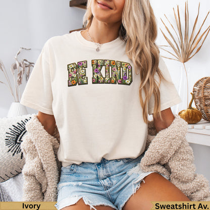 Cream t-shirt with "BE KIND" in floral block letters, worn by a woman.