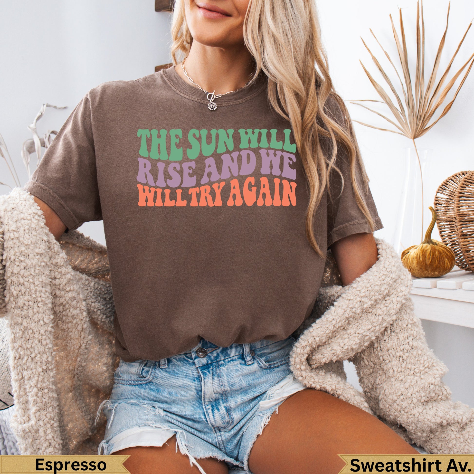 Brown sweatshirt with colorful "The sun will rise and we will try again" text.