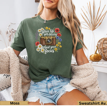 Green t-shirt with camera graphic and floral border, text reads "when life gets blurry adjust your focus".