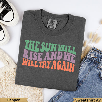 Dark grey t-shirt with "The Sun Will Rise And We Will Try Again" in colorful text.