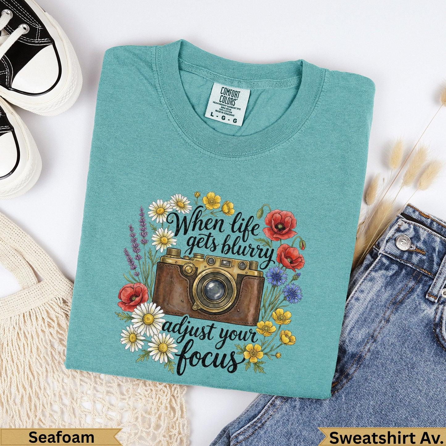 Teal graphic t-shirt with vintage camera and floral design, text "When life gets blurry adjust your focus".