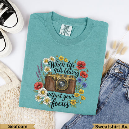 Teal graphic t-shirt with vintage camera and floral design, text "When life gets blurry adjust your focus".