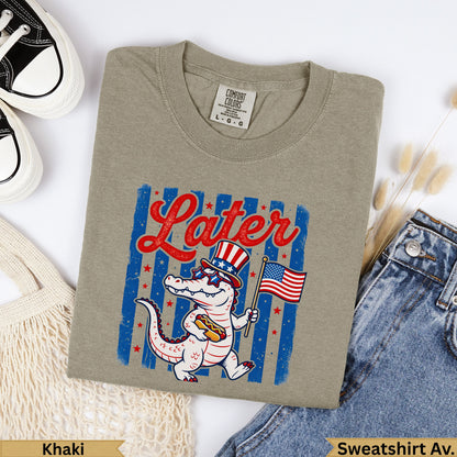 Khaki crewneck sweatshirt with alligator wearing sunglasses and holding hot dog with American flag.