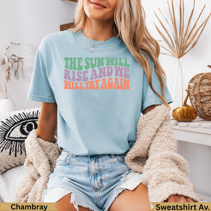 Light blue t-shirt with colorful text "The sun will rise and we will try again."