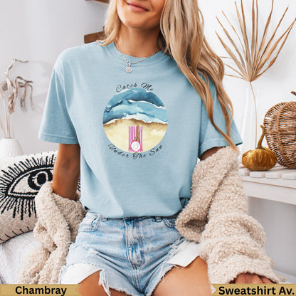 Light blue t-shirt with beach scene and "Catch Me Under The Sun" text.