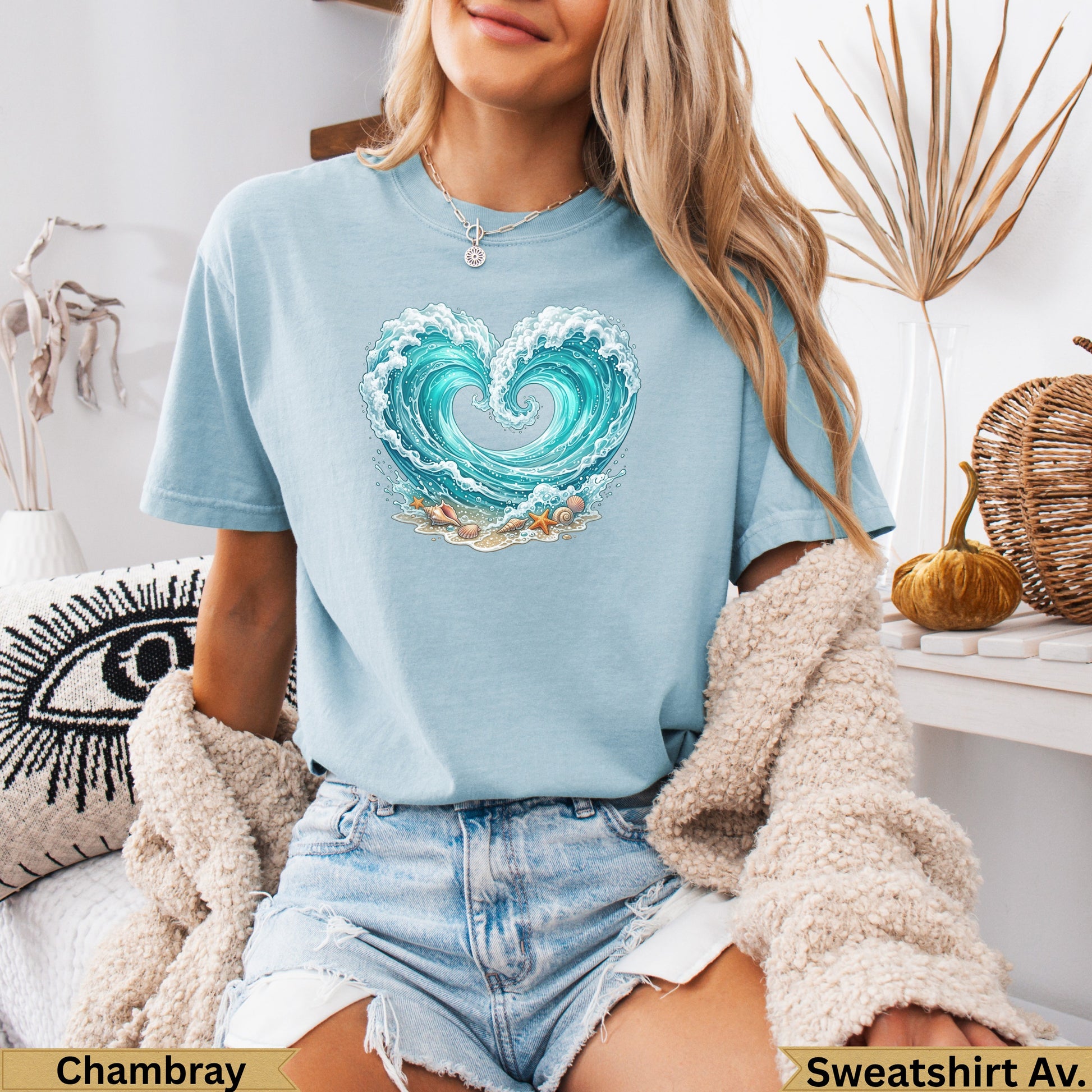 Light blue t-shirt with heart-shaped wave graphic and seashells on sand.