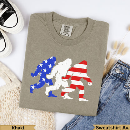 Three Bigfoot figures in US flag patterns on a khaki t-shirt.